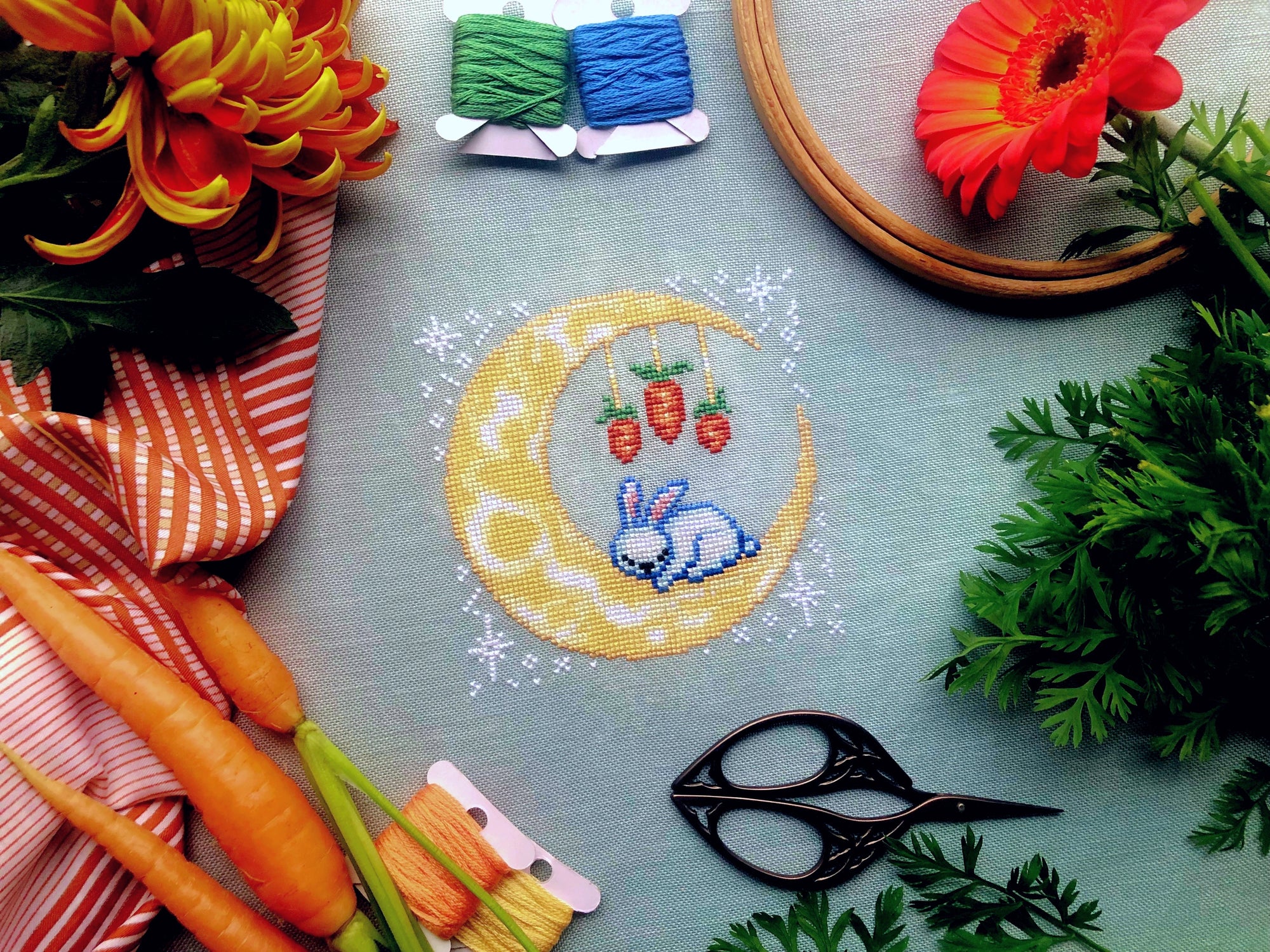 Front view of Dreaming of Carrots cross stitch pattern. Scene is surrounded by flowers, carrots and embroidery supplies. Moon is round but is surrounded by white stars that create a square composition. Bunny looks peaceful and content.