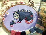 Front view of possum mama cross stitch pattern. The opossum mother has 6 babies, 5 of which are on her back. The 6th one is sitting on her left. Colors are mainly blueish grey, lilac, beige and pink. The baby possums are extremely cute.