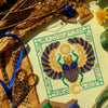 Top down, angled view of Egyptian Scarab cross stitch pattern by NeedleLot Designs. Colors are blue, old gold, green, black and yellow. Piece consists only of DMC threads. Scarab is flanked by sun and moon, and is surrounded by hieroglyphs.