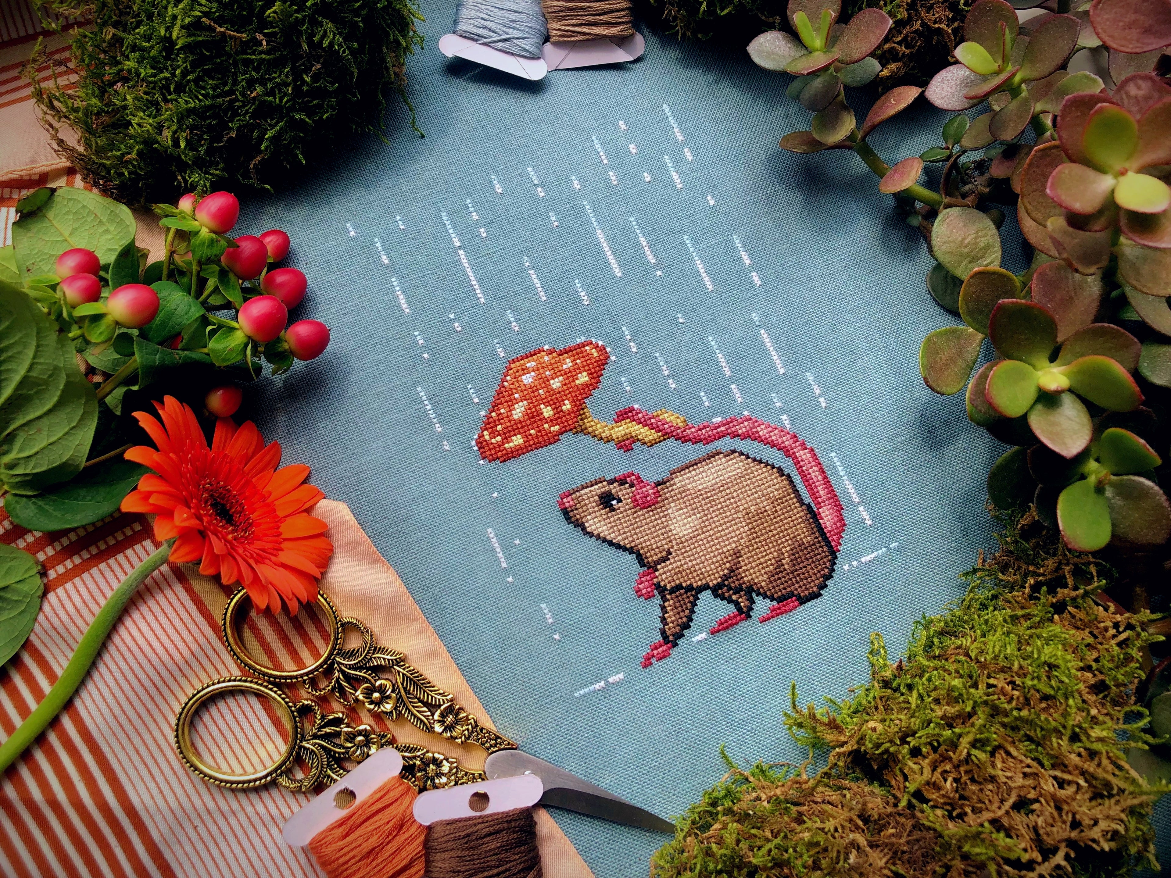 Topdown image of Rainy days rat cross stitch pattern. Fabric is turqois-blue, rat is brown with a pink tail. He is holding a mushroom over his head. His head is tilted upwards. His little paw is in the air. The mushroom keeps the rain out of his face
