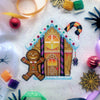 Flat-lay of Cookies & Scream gingerbread house cross stitch pattern. Finished piece is of medium size. Colors are yellow, brown, orange, purple, black and white. Gingerbread man is standing in front of gingerbread house with a Halloween smile.