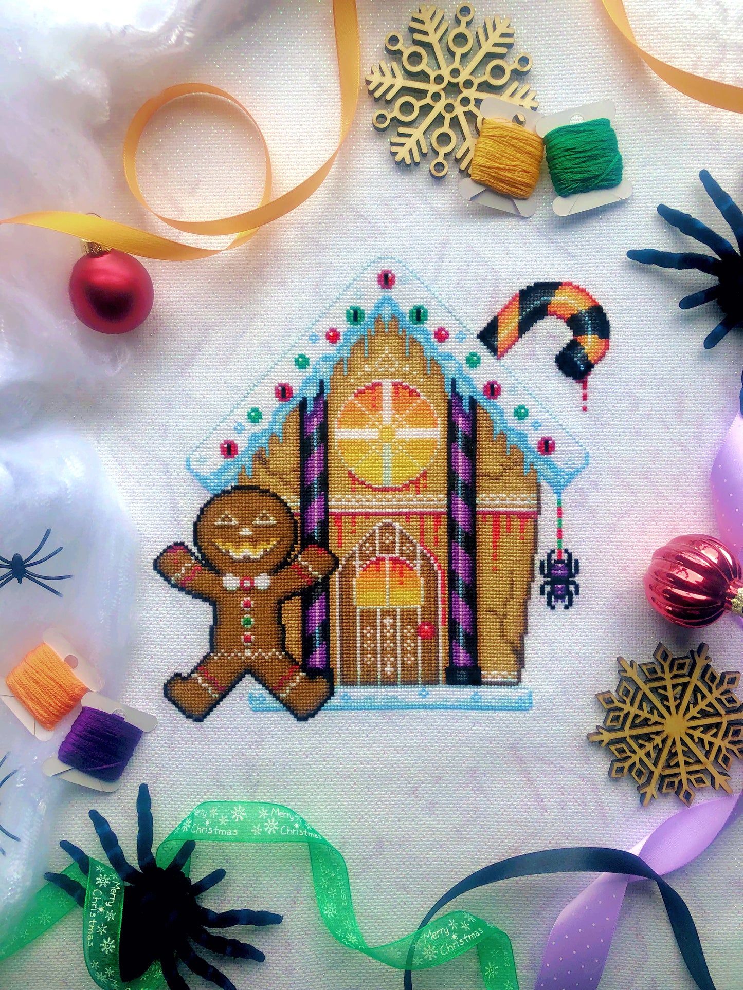 Flat-lay of Cookies & Scream gingerbread house cross stitch pattern. Finished piece is of medium size. Colors are yellow, brown, orange, purple, black and white. Gingerbread man is standing in front of gingerbread house with a Halloween smile.