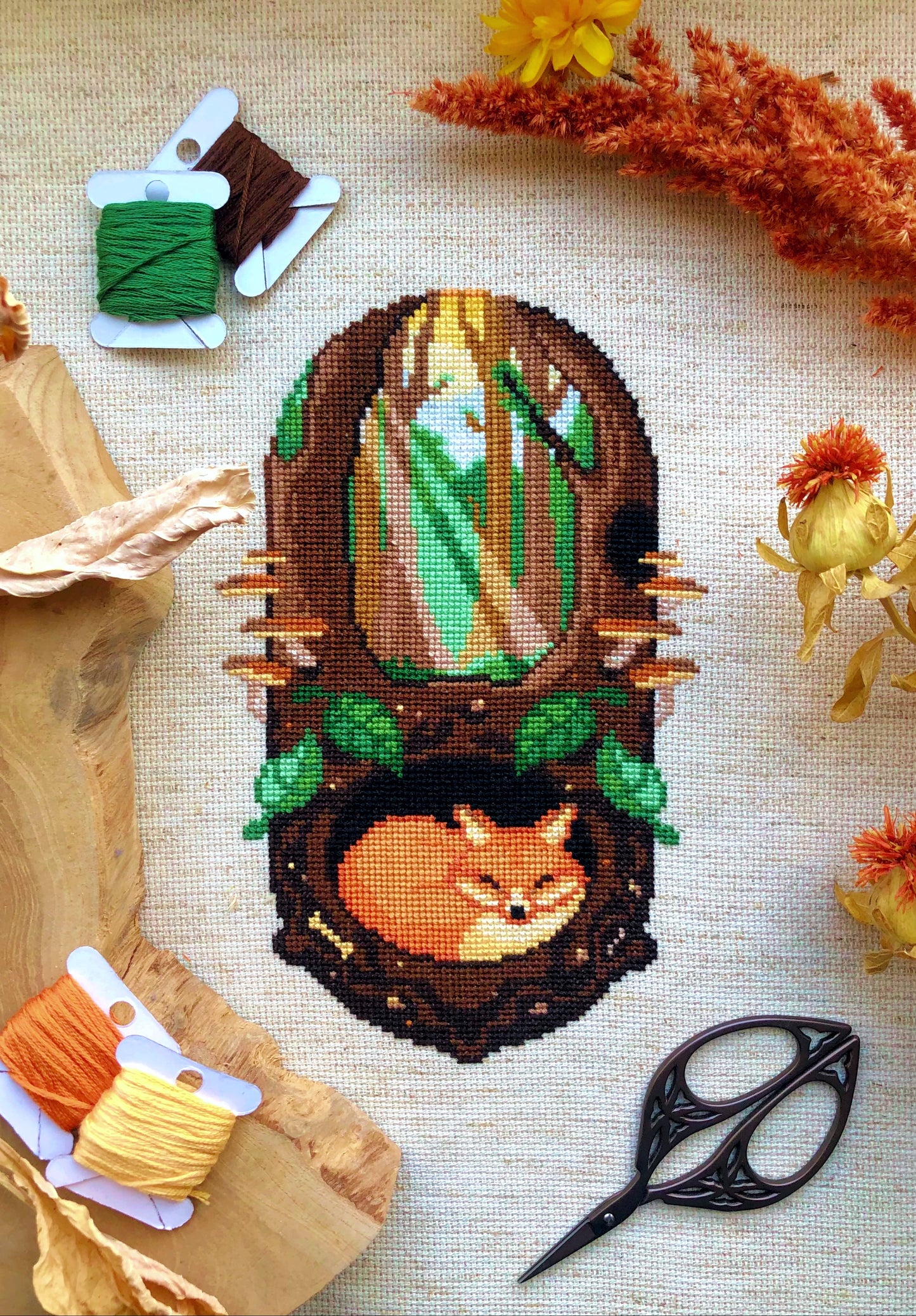 Vertical flat-lay of Sleeping Fox cross stitch pattern. Stitched item, surrounded by decorations. Finished piece is of medium size. Colors are green, brown, orange, black, beige and white. Fox is sleeping underground. Trees and sun rays are above.
