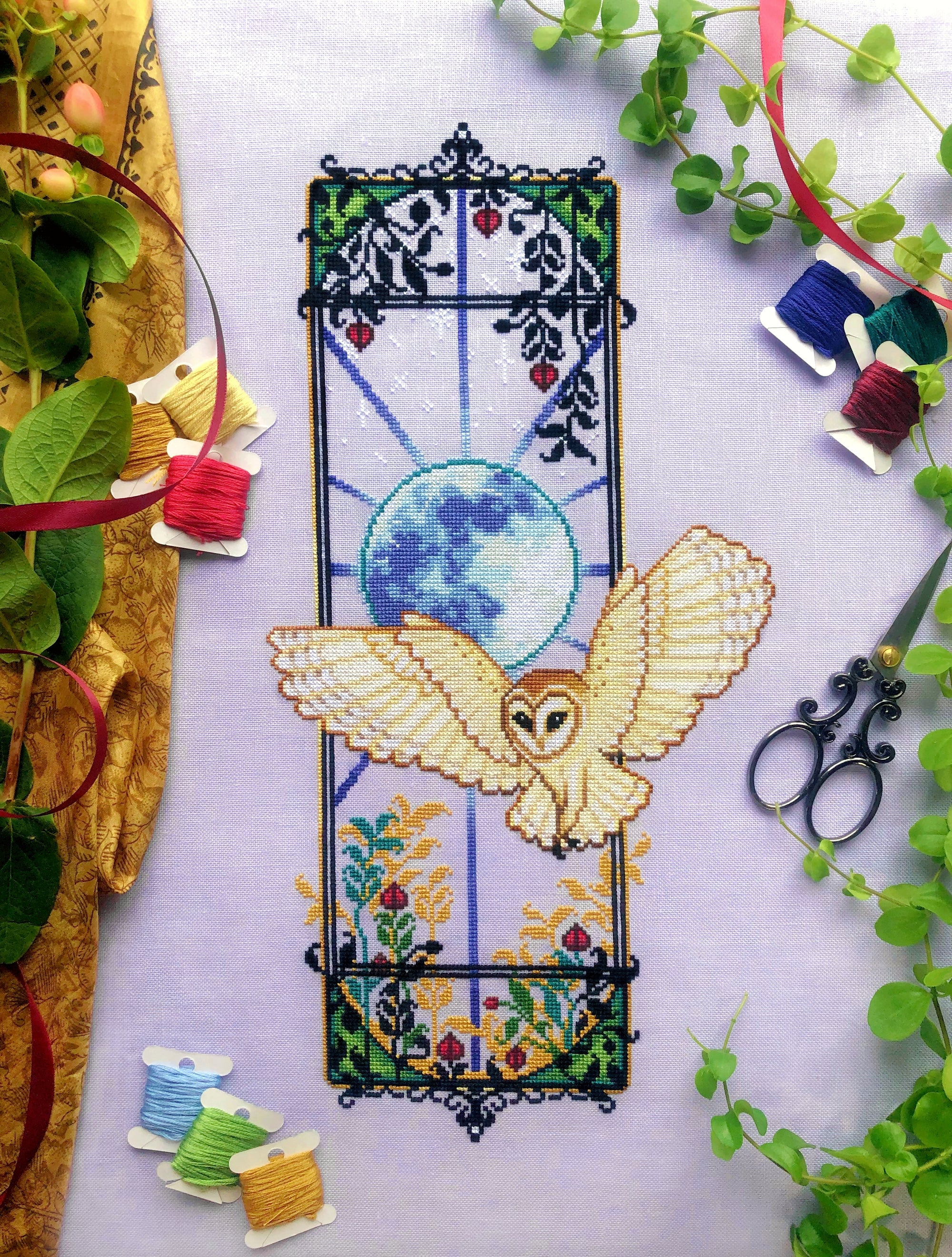 Front view of barn owl Jugendstil cross stitch pattern. The piece is large and vertical. The owl moves outside of the boundaries of the window. There are lots of plants surrounding the scene. Colors are mainly black, blue, green, teal, red, beige.