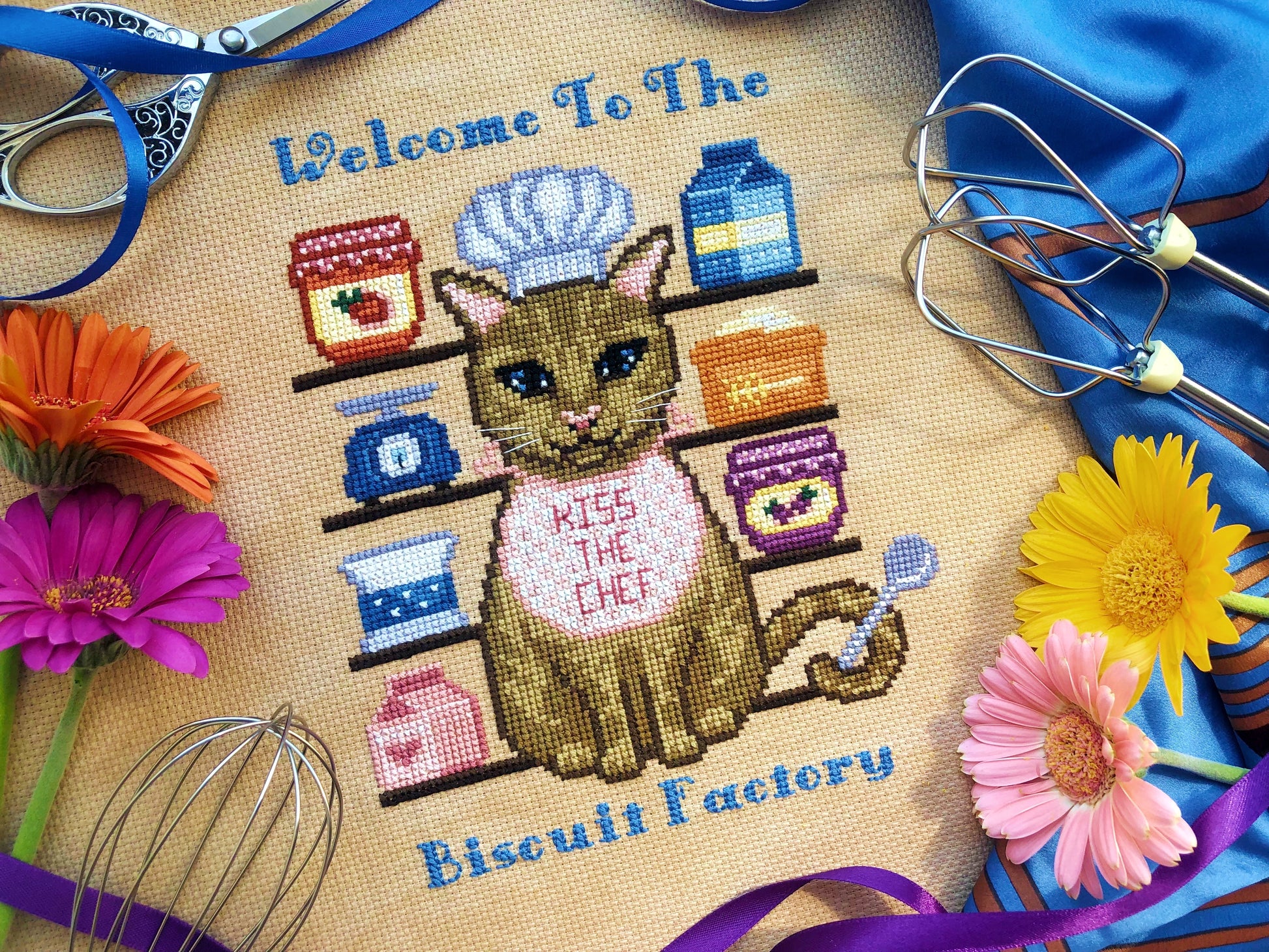 Front view of cat baker. Composition consists of a tabby cat, surrounded by baking supplies such as jams, milks, sugar and wheat. The cat is holding a spoon with his tail. The text positioned above and below reads "Welcome to the Biscuit Factory".