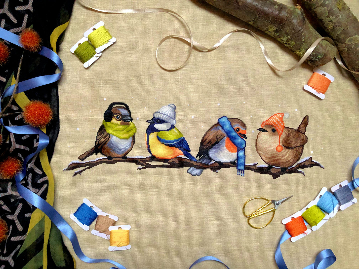 Flatlay of Winter Birds cross stitch pattern. Stitched item is surrounded by decorations. Finished piece is of medium to large size. Colors are yellow, brown, orange, blue, green, white. There are four birds sitting on a branch wearing winter gear.