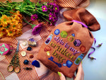 Trinket bag cross stitch front view. The phrase This is my Trinket Bag is stitched onto a drawstring bag. It is surrounded by objects, such as dice, potion bottles, mushrooms, buttons, a key and a ring. The bag is brown. The scene is very colorful.