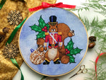 Front view of Nutcracker squirrel cross stitch pattern. Squirrel is wearing a military suit and looks stern. His jacket is red and he is wearing black boots and a hat. Around him are walnuts, acorns, holly and a bauble. Scene is surrounded by decor.