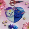 Front view of Sun and Moon Cats cross stitch pattern. Cats are snuggled together in a heart-shaped frame, with two different sky backgrounds, one containing clouds and the other containing stars. Cats have a sun and moon on their forehead.