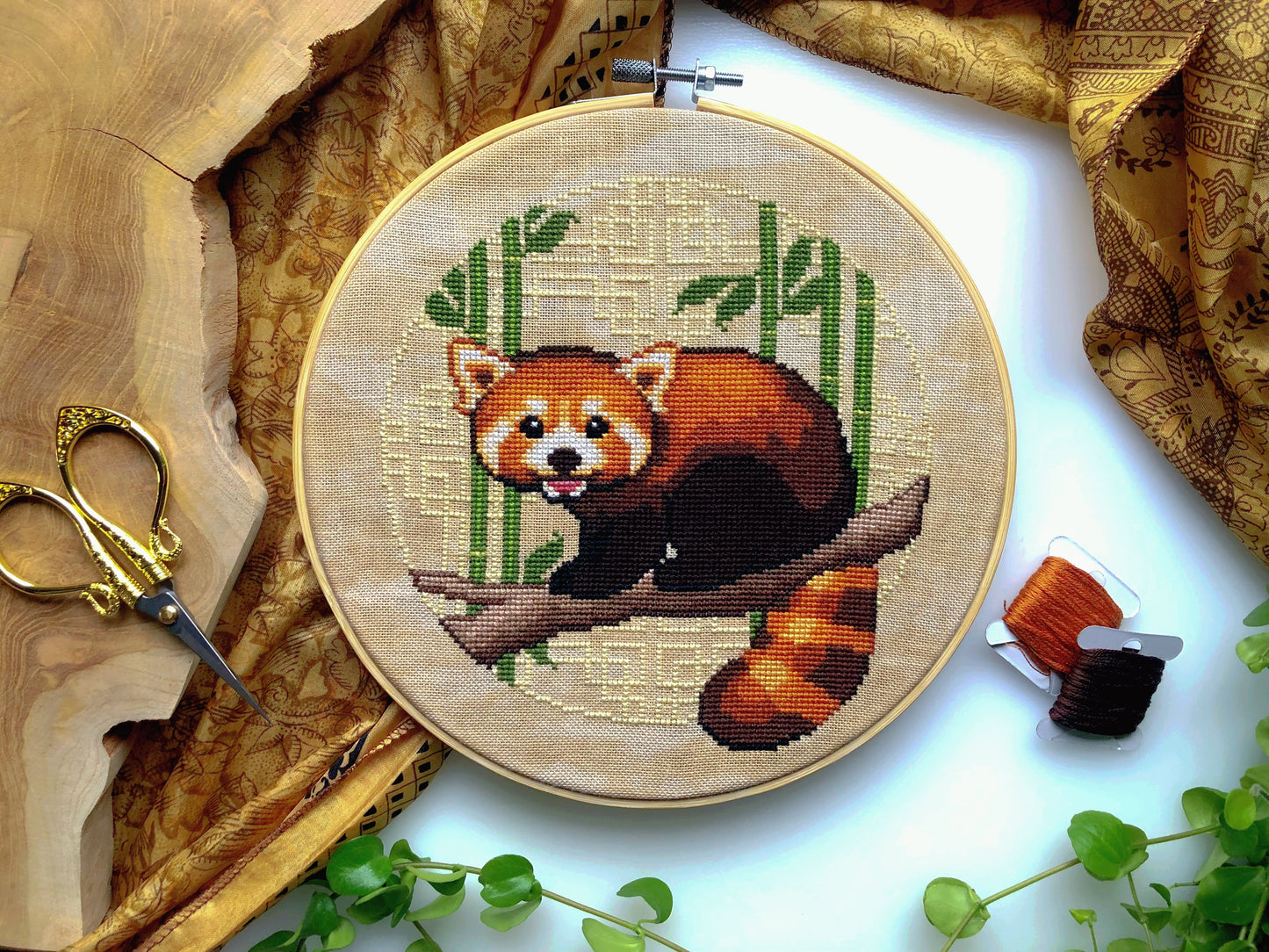 Flat-lay of Red Panda cross stitch pattern. Stitched item, surrounded by foliage, scissors and decor. Finished piece is of small to medium size. Colors are yellow, brown, green and white. Red Panda is cute and small and is looking at the viewer.