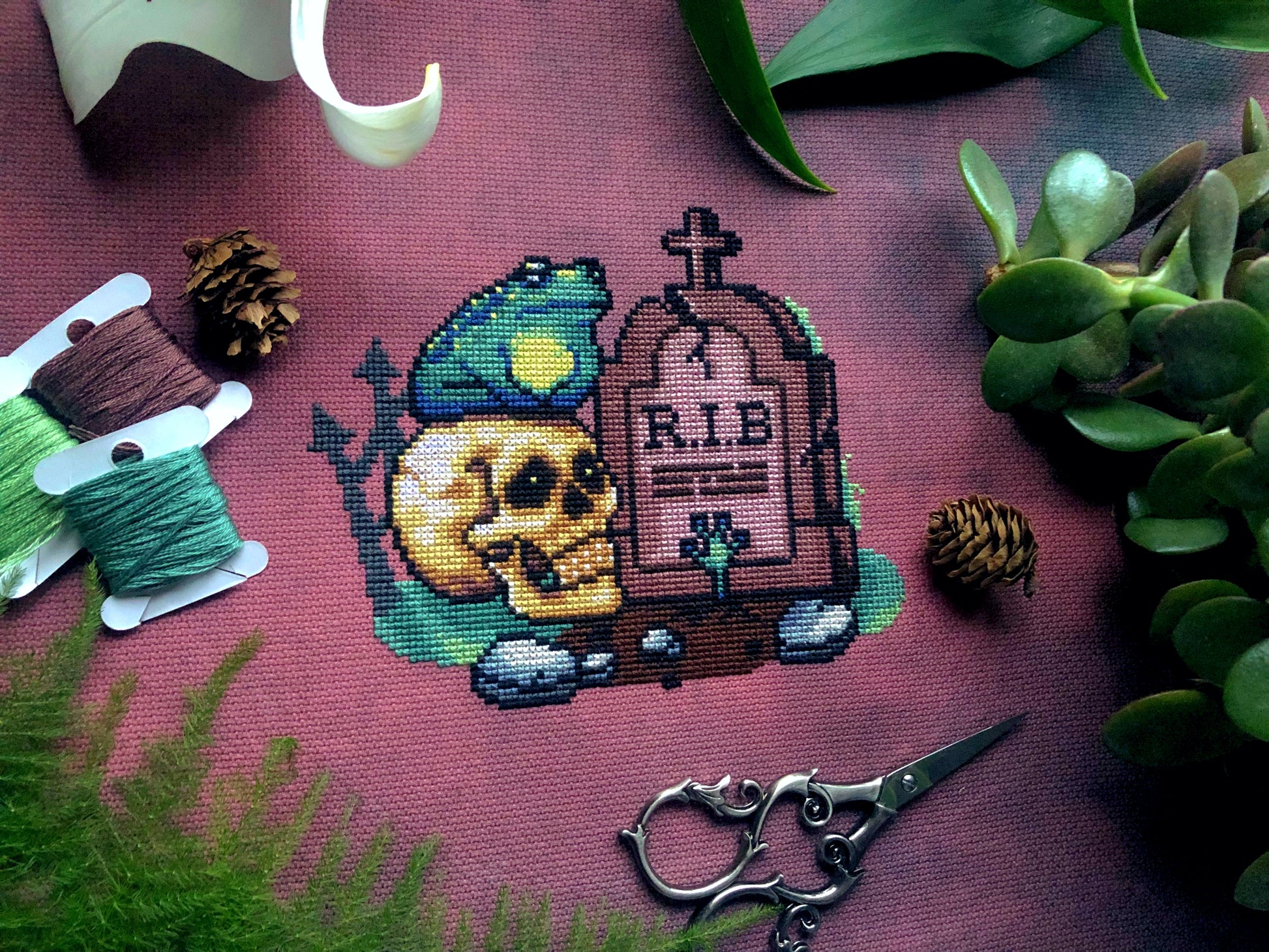 Front view of frog graveyard Rest in Bog cross stitch pattern. Frog is sitting on a realistic looking skull, next to a tombstone that says R.I.B. Fabric is a brown/purple shade. Scene is surrounded by foliage, scissors, pinecones and bobbins.