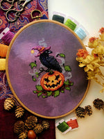 Flat-lay of Jack-Crow-Lantern cross stitch pattern. Stitched item, surrounded by decorations. Finished piece is small. Colors are orange, purple, black, and green. Crow is wearing a hat and looking over its shoulder, standing on a smiling pumpkin.