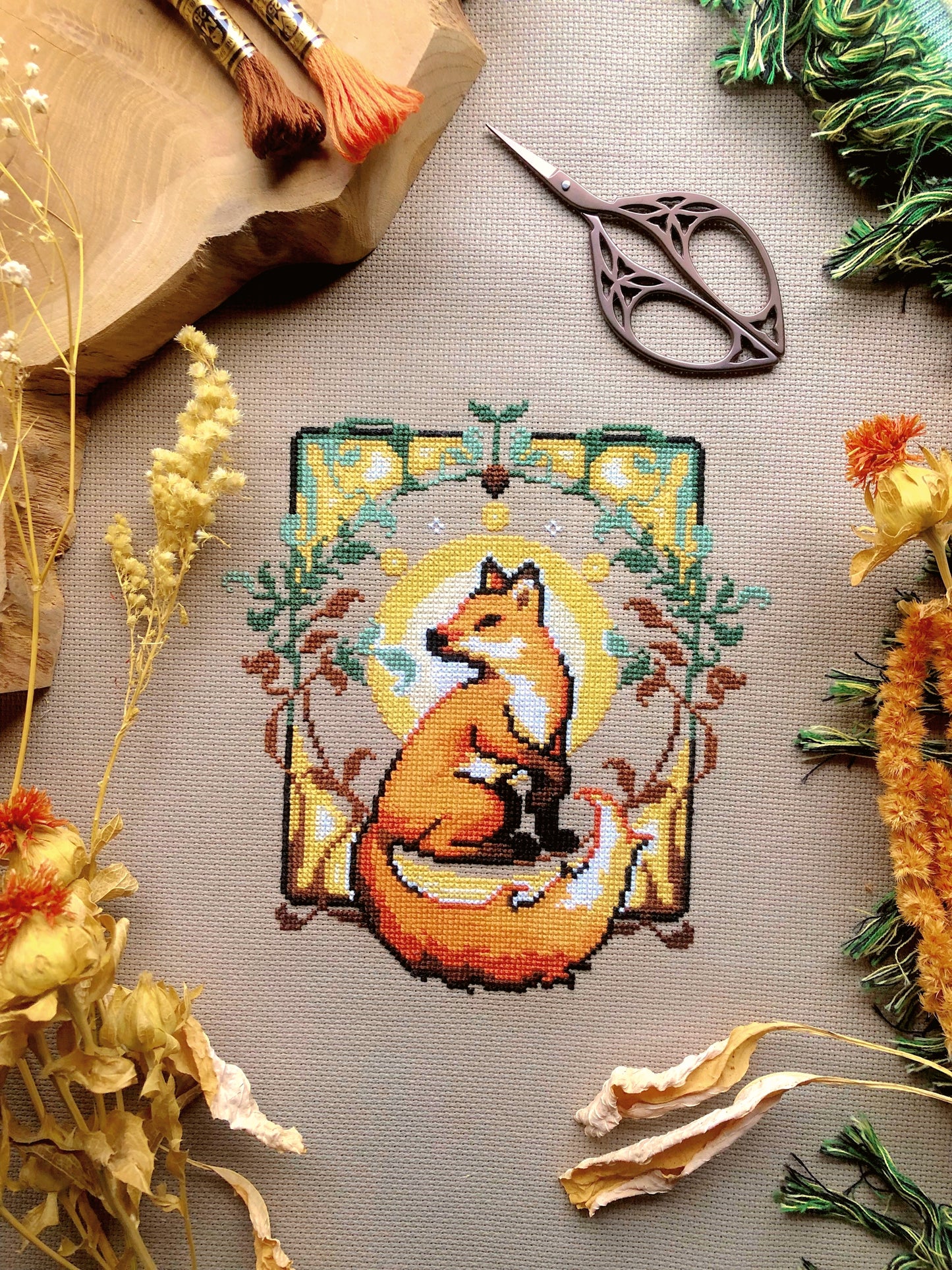 Flat-lay of Art Nouveau Fox cross stitch pattern. Stitched item, surrounded by decorations. Finished piece is of small to medium size. Colors are yellow, brown, orange, green and white. Fox is cute and small and is looking over its shoulder.