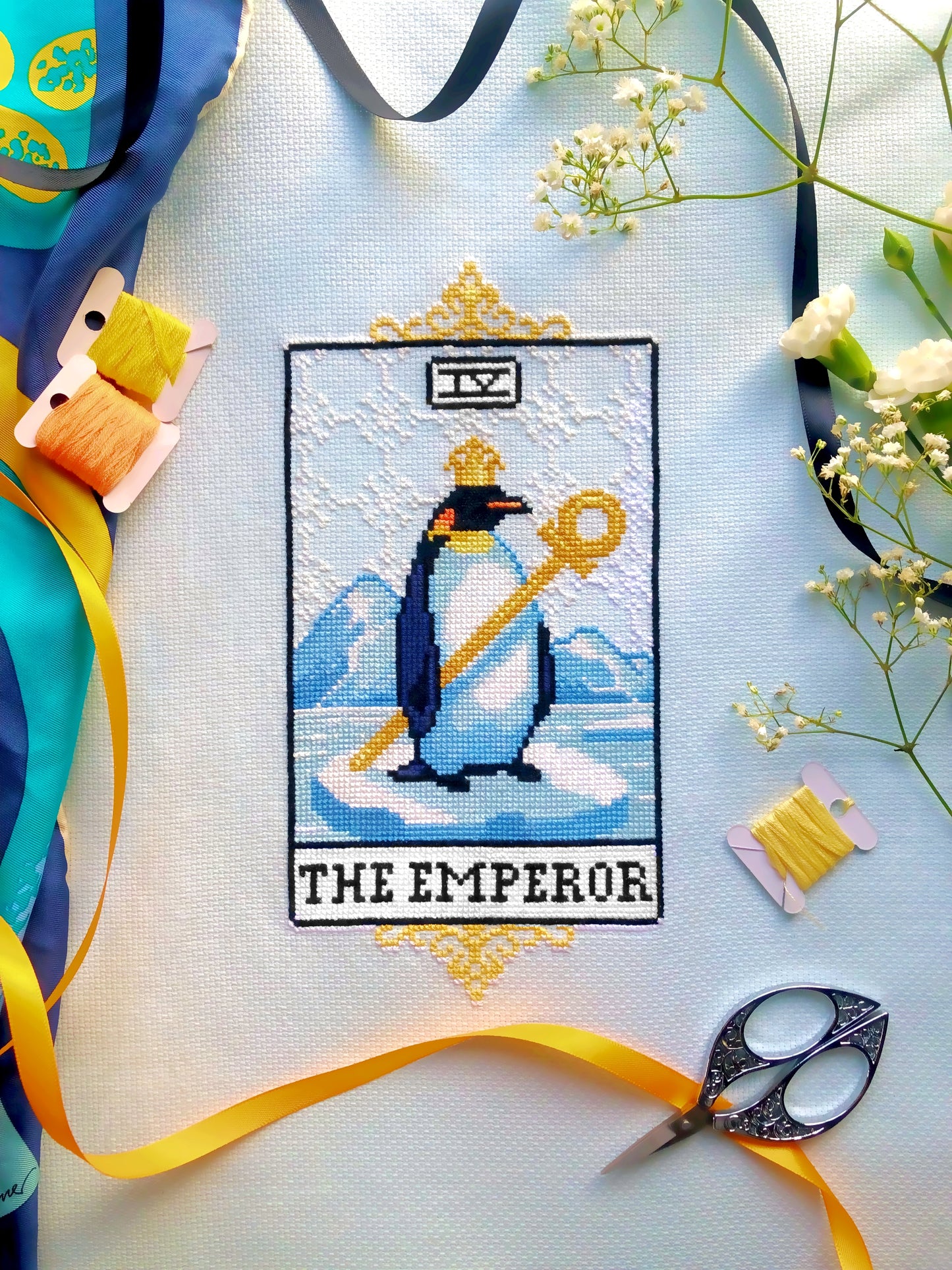 Flat-lay of The Emperor tarot cross stitch pattern. Stitched item, surrounded by decorations. Finished piece is of small size. Colors are yellow, blue, black, white and orange. Emperor penguin is holding a sceptor and wearing a crown.