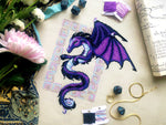 Flat-lay of Dragon cross stitch pattern. Stitched item, surrounded by decorations. Finished piece is of medium to large size. Colors are blue, purple, white, black and pink. Dragon is surrounded by variegated frame. Its head is like that of a horse.