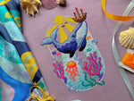Font view of Whale with Boat cross stitch pattern. Blue whale has a boat on its nose that it has lifted out of the water. The sun is shining. Underwater we can see coral, fish, seaweed and a jellyfish. Colors are very vibrant. Birds are in the sky.