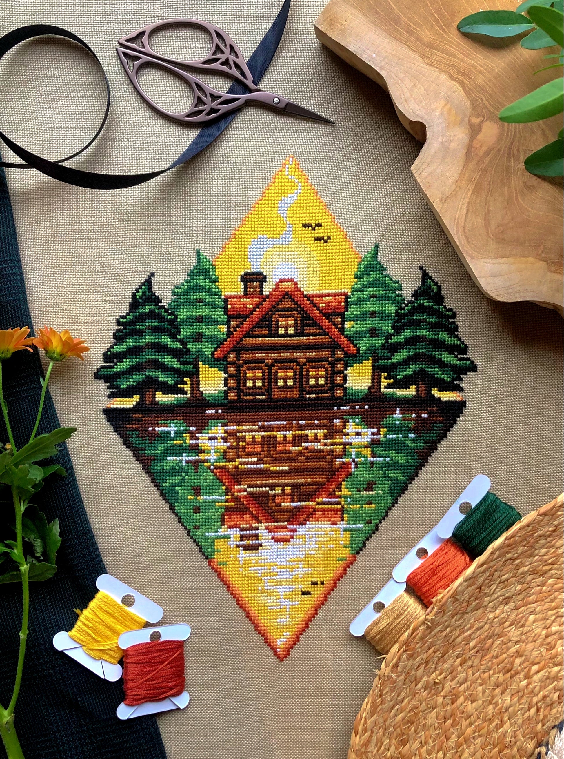 Flat-lay of Lakeside Cabin cross stitch pattern. Stitched item, surrounded by decorations. Finished piece is of large size. Colors are yellow, brown, orange, green, black and white. Scenery contains a sunset, and the cabin is reflected in the water.