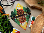 Flat-lay of Lakeside Cabin cross stitch pattern. Stitched item, surrounded by decorations. Finished piece is of large size. Colors are yellow, brown, orange, green, black and white. Scenery contains a sunset, and the cabin is reflected in the water.