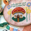 Closer view of toadstool mushroom house cross stitch pattern. House is framed on either side by yellow and lilac flowers. The house has a little round door and several windows. In front is a path, surrounded by green grass. There is a lantern.
