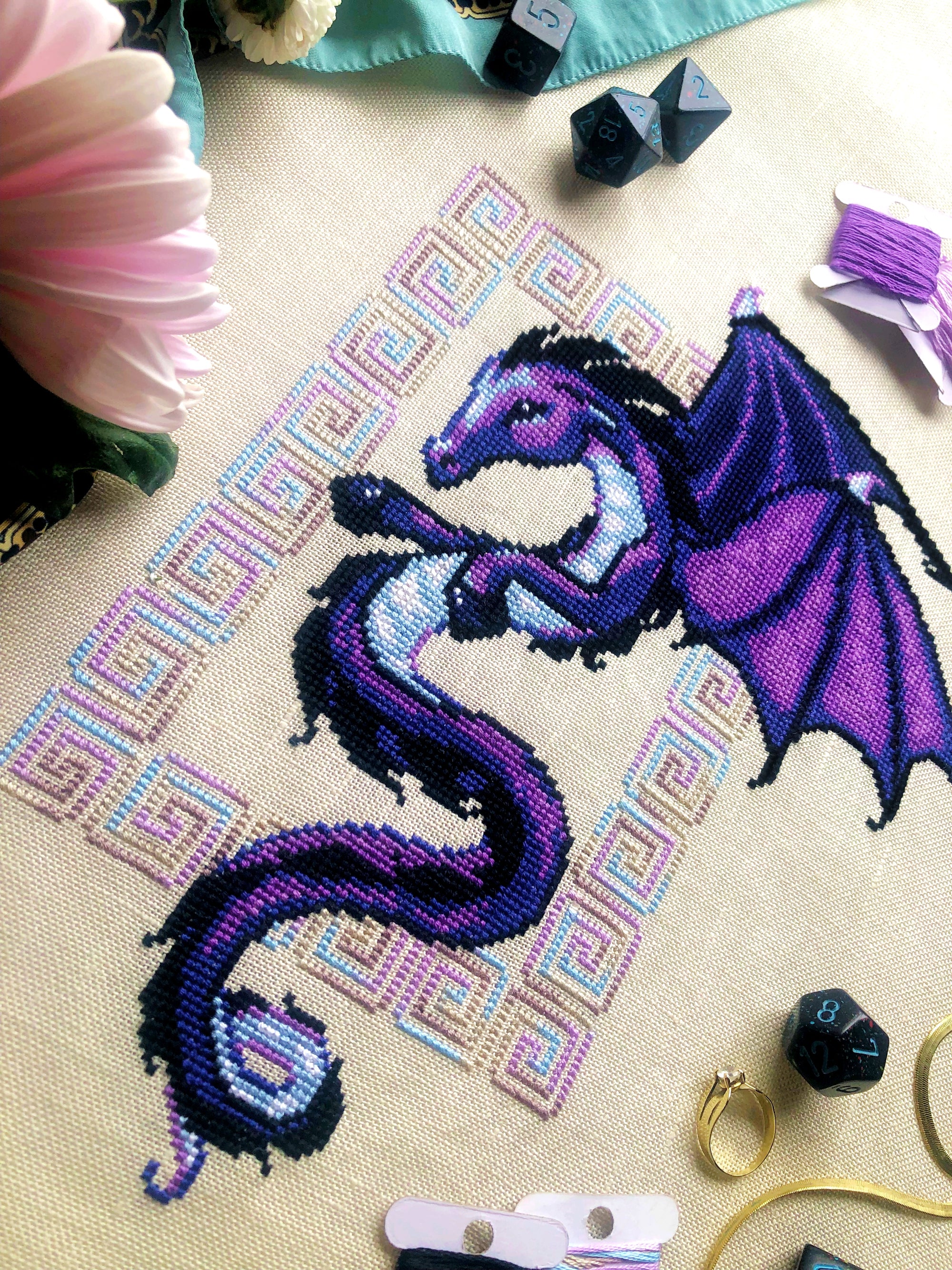 Flat-lay of Dragon cross stitch pattern. Stitched item, surrounded by decorations. Finished piece is of medium to large size. Colors are blue, purple, white, black and pink. Dragon is surrounded by variegated frame. Its head is like that of a horse.