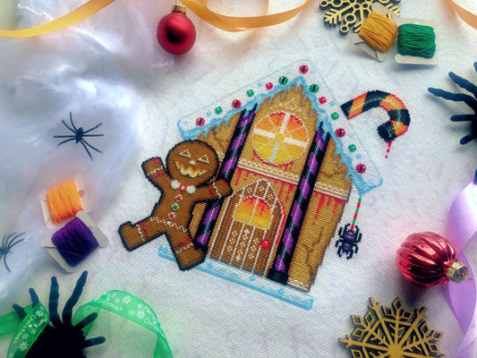 Flat-lay of Cookies & Scream gingerbread house cross stitch pattern, at a slight angle. Finished piece is of medium size. Piece is bright and colorful, eminating both Christmas spirit and Halloween spirit. Candy and candycanes are everywhere.