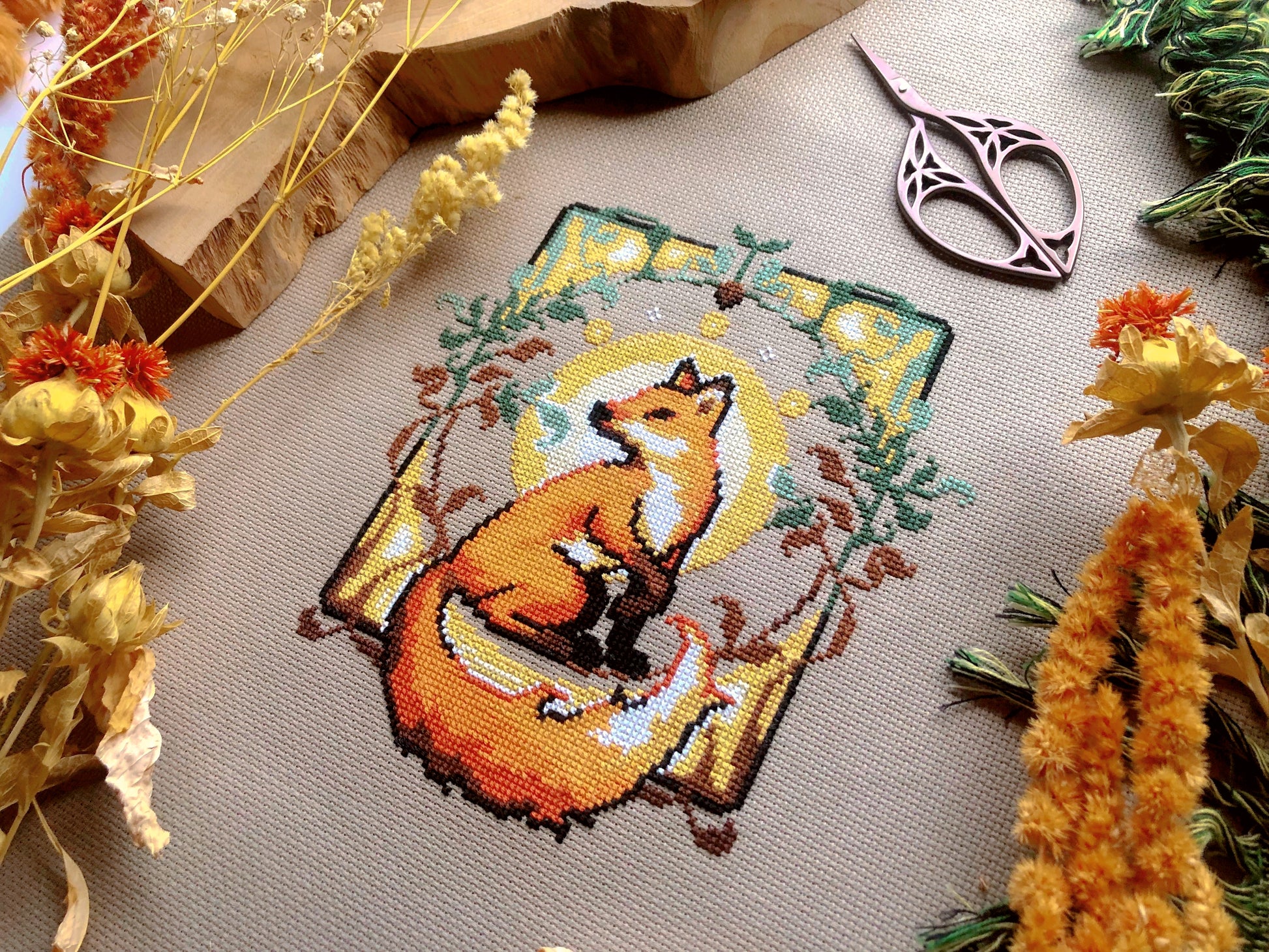Flat-lay of Art Nouveau Fox cross stitch pattern. Stitched item, surrounded by decorations. Finished piece is of small to medium size. Colors are yellow, brown, orange, green and white. Fox is cute and small and is looking over its shoulder.