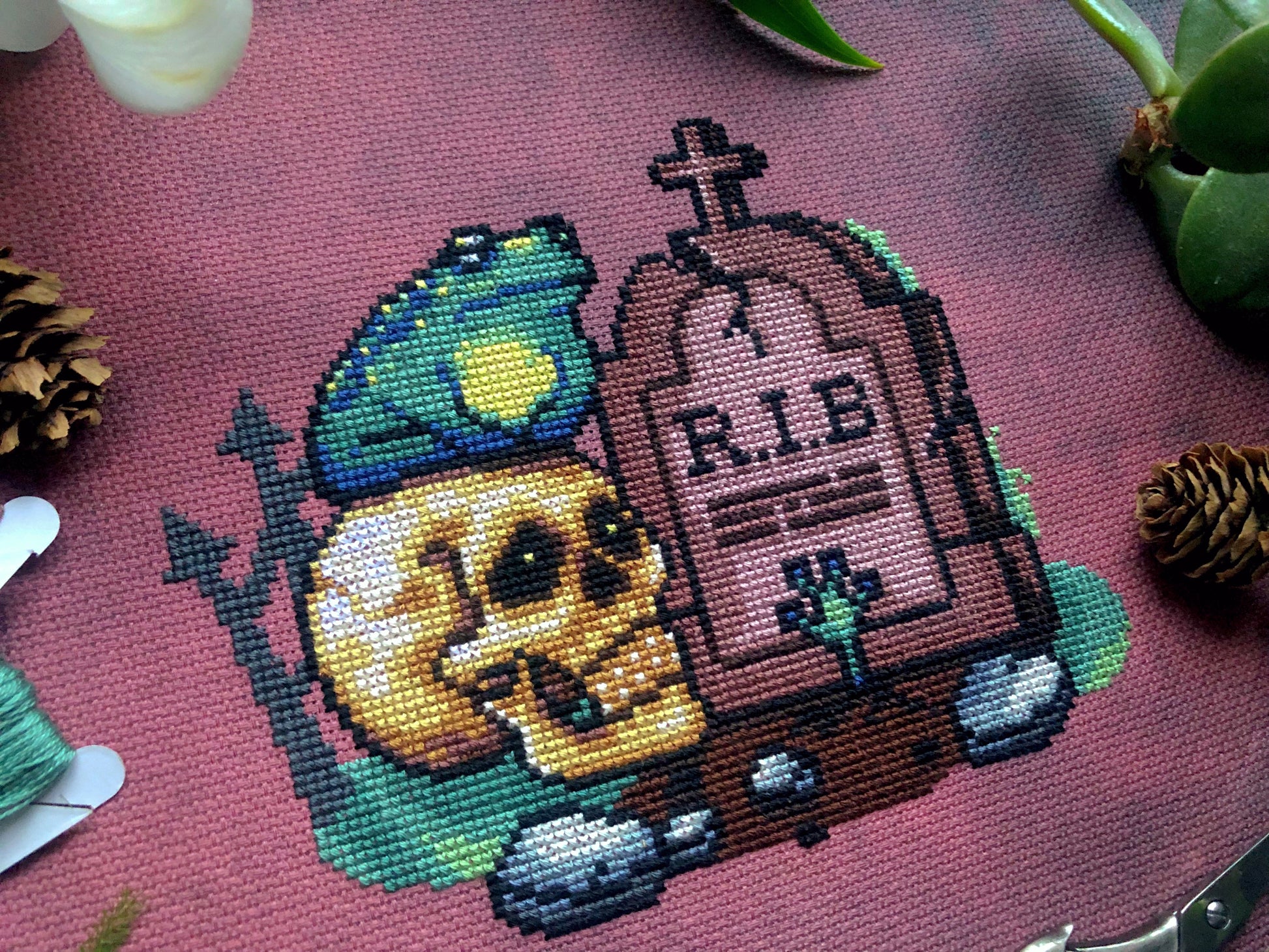 Closeup of frog graveyard Rest in Bog cross stitch pattern. Colors are vibrant. Frog is sitting on a realistic looking skull, next to a tombstone that says R.I.B. From the grave itself a frog hand sticks up, like that of a zombie.