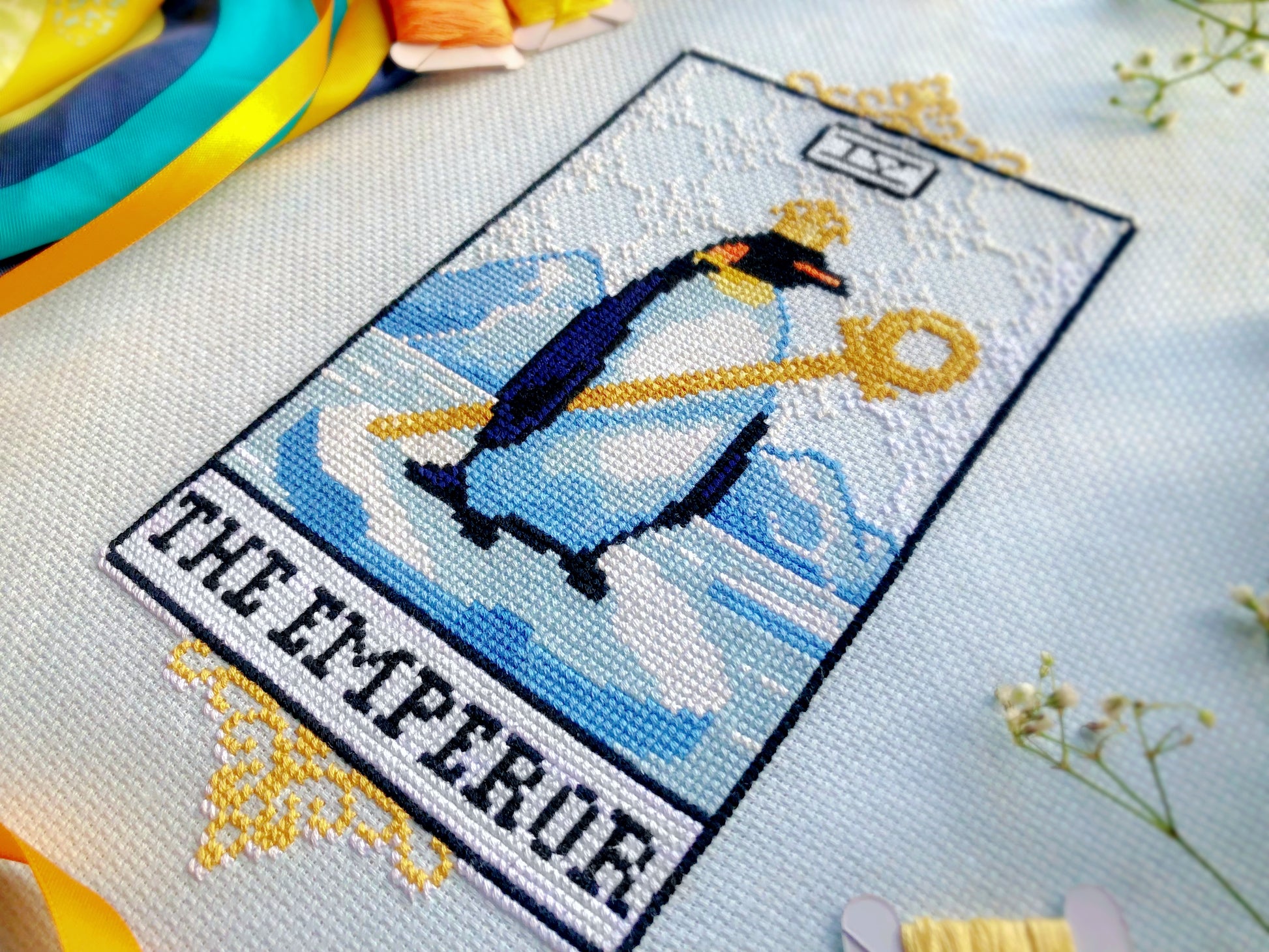Closeup of the Emperor tarot cross stitch pattern. Image is focused on the lower right corner of the design. Emperor penguin is standing proudly in his attire on a block of ice, surrounded by water and icebergs. The frame is that of a tarot card.