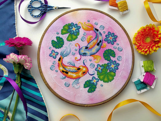 Flat-lay of Koi Fish cross stitch pattern. Stitched item, surrounded by decorations. Finished piece is of medium size. Colors are very bright, and are green, blue, orange, pink and yellow. There are two fish, surrounded by lotus leaves and flowers.