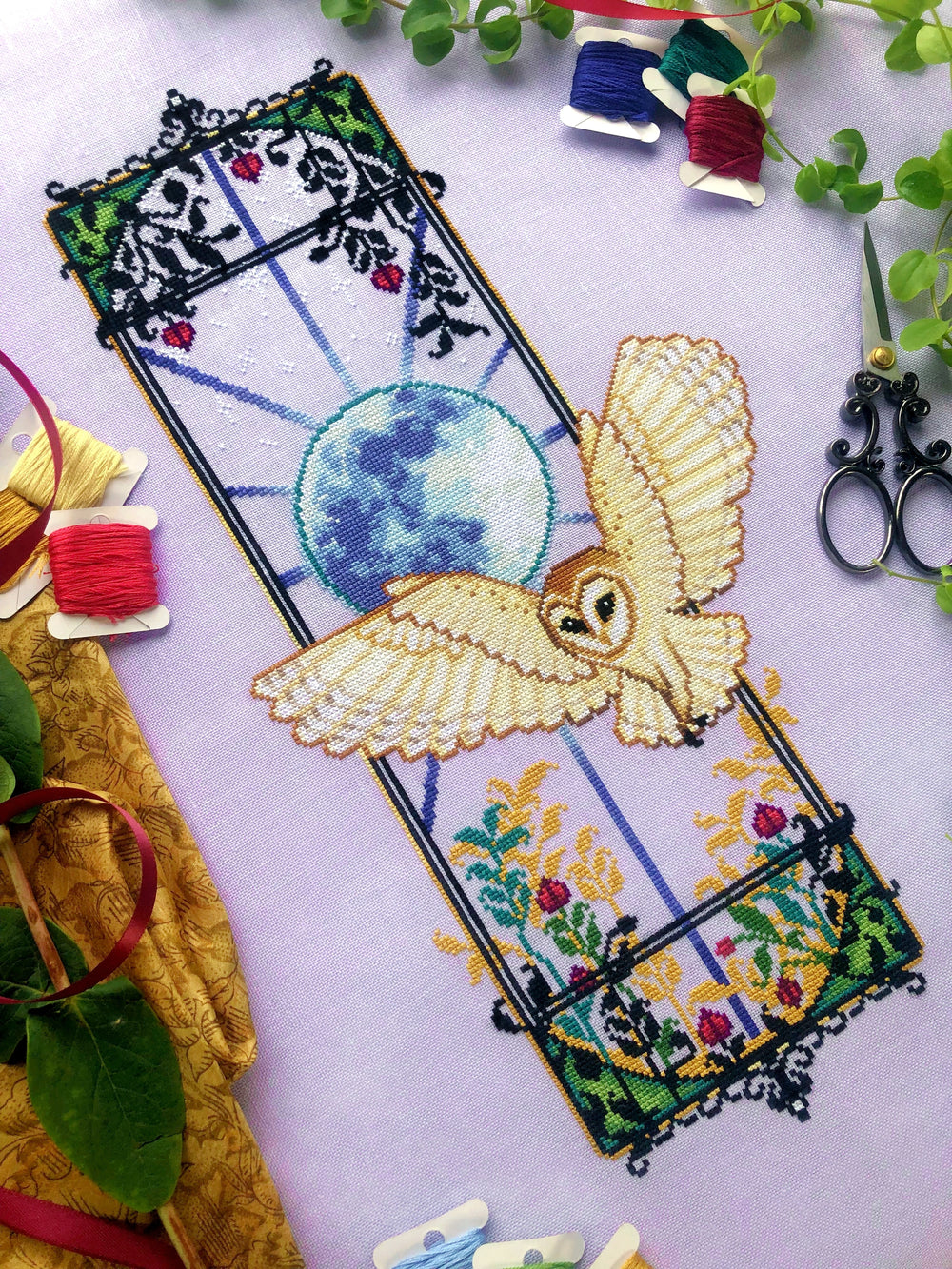 Angled front view of Art Nouveau barn owl cross stitch pattern. Finished piece is elongated and looks like a window. There is a big blue moon in the middle that radiates outward. A barn owl is flying into the scene with its wings spread.