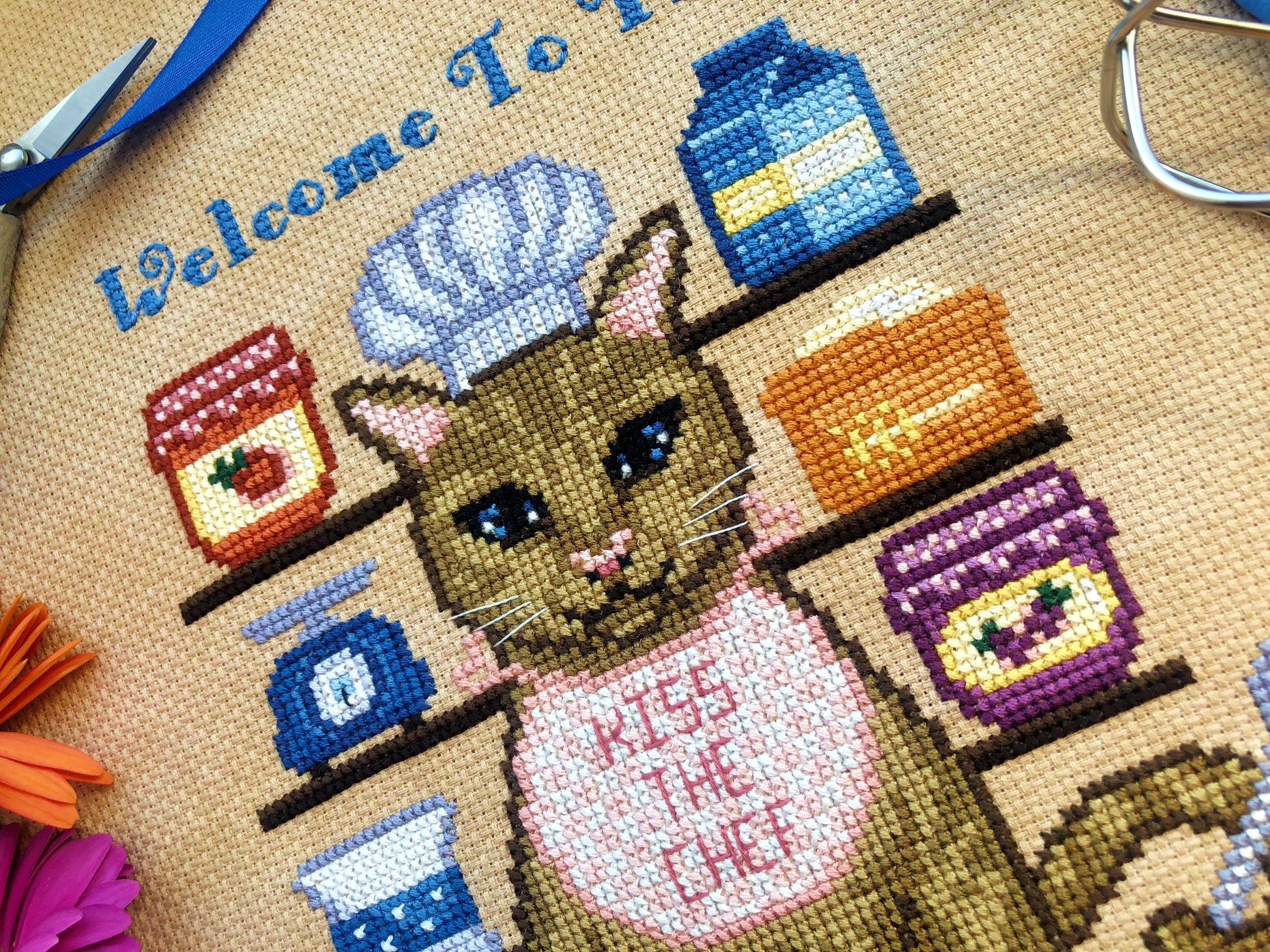 Closeup of cat baker. He is wearing a white chef's hat, and is wearing a checkered apron that says "Kiss the Chef". Colors are orange, brown, pink, purple, red, yellow and blue. Cat's eyes are blue, and he has whiskers. Stitches are neat and tidy.