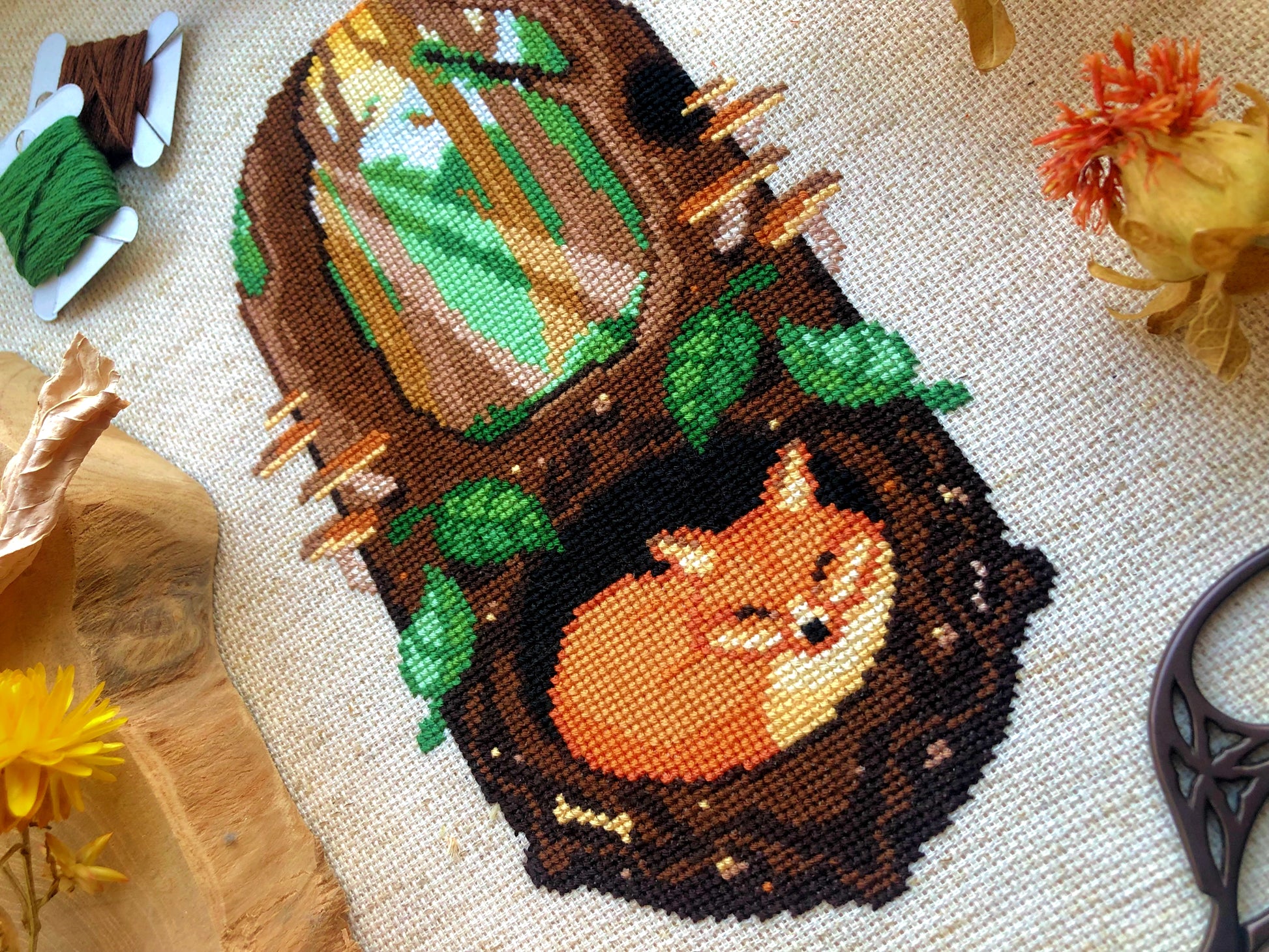 Closeup of Sleeping Fox cross stitch pattern. Stitched item, surrounded by decorations. Finished piece is of medium size. Colors are green, brown, orange, black, beige and white. Fox is sleeping underground. Trees and sun rays are above.