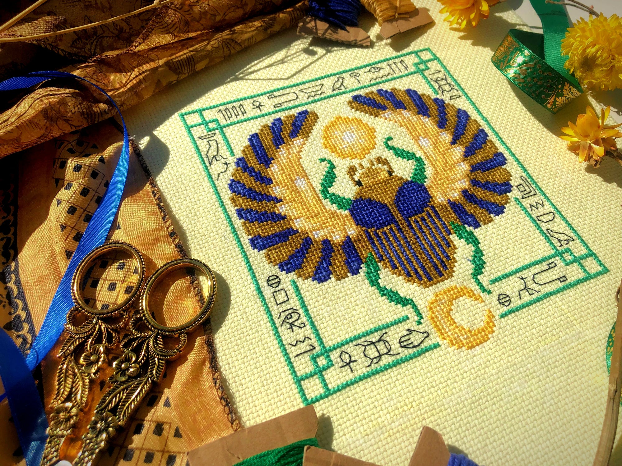 Top down, angled view of Egyptian Scarab cross stitch pattern by NeedleLot Designs. Colors are blue, old gold, green, black and yellow. Piece consists only of DMC threads. Scarab is flanked by sun and moon, and is surrounded by hieroglyphs.