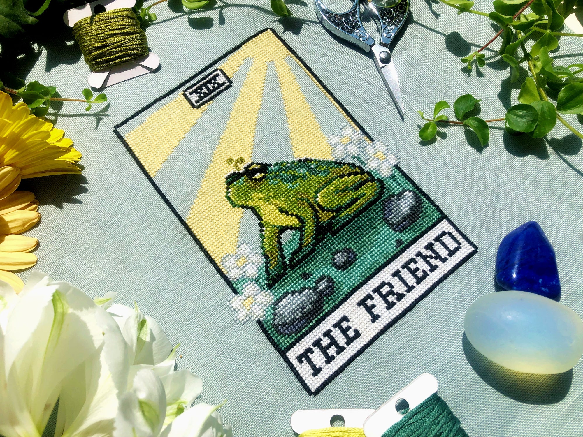 Flatlay of The Friend frog tarot cross stitch pattern. Stitched item, surrounded by decorations. Finished piece is of small to medium size. Colors are yellow, green, blue, grey and black. Frog is cute and small. The Friend is written underneath.