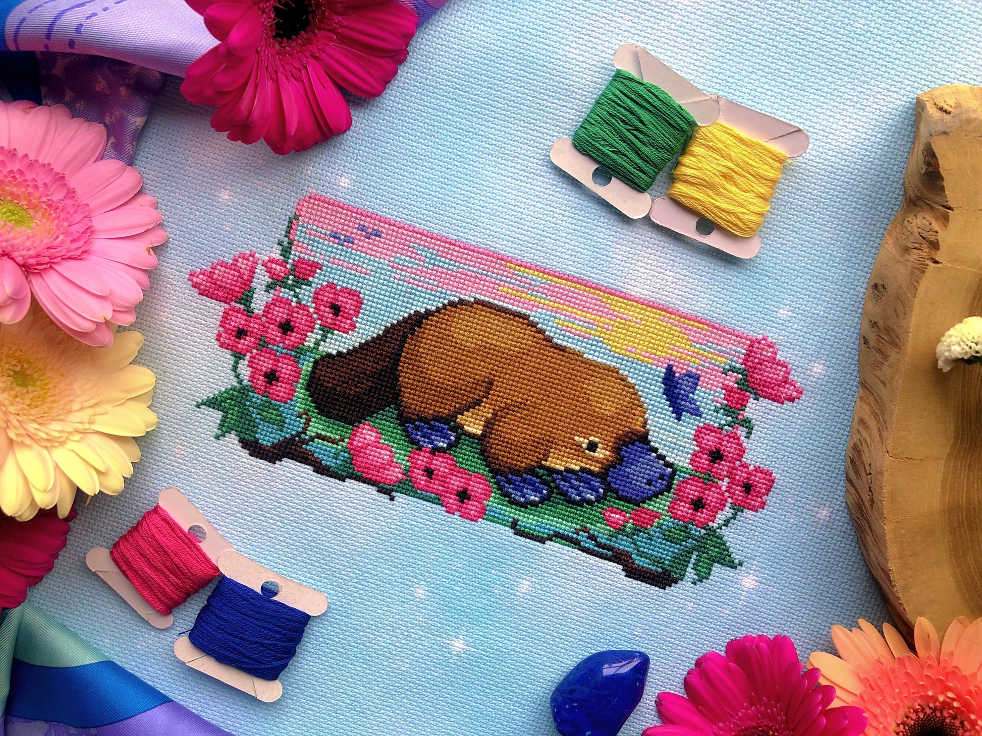 Platypus in a flower field cross stitch pattern, surrounded by flowers and bobbins. Colors are bright. Mainly pink, blue, yellow, green, and brown are present. Platypus is facing right, and is surrounded by pink flowers. There is a blue butterfly.