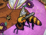 Closeup view of Queen Bee cross stitch pattern. Underneath the Queen Bee is an actual bee, of much smaller size. The bee is flying and is yellow with black brown stripes. Colors are vibrant and stitches are neat and tidy. Queen's skin is a deep brown