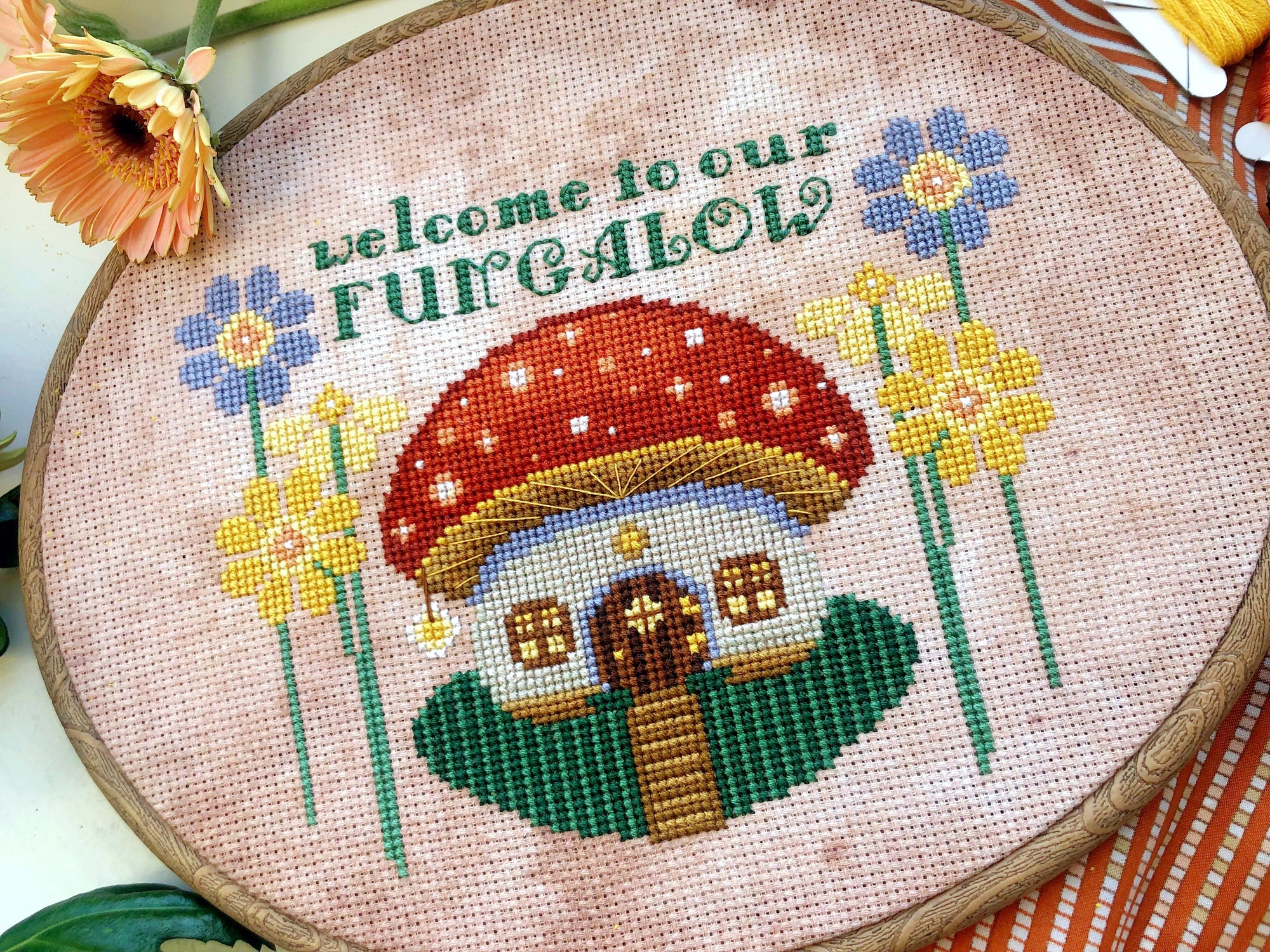 Closer view of toadstool mushroom house cross stitch pattern. House is framed on either side by yellow and lilac flowers. The house has a little round door and several windows. In front is a path, surrounded by green grass. There is a lantern.