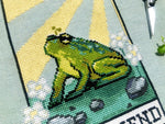 Closeup of The Friend frog tarot cross stitch pattern. Stitched item, surrounded by decorations. Finished piece is of small to medium size. Colors are yellow, green, blue, grey and black. Frog is cute and small. The Friend is written underneath.