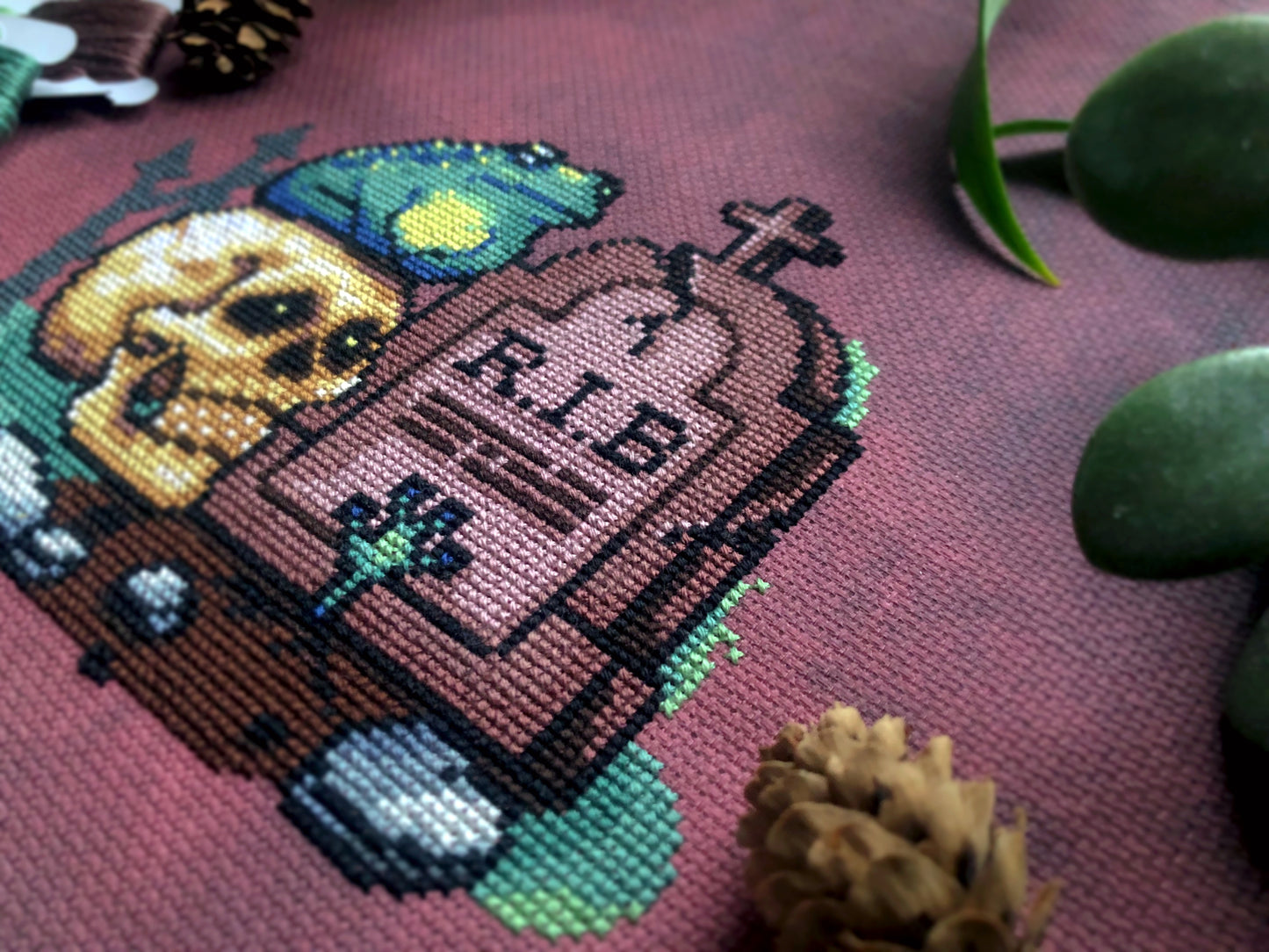 Closeup of R.I.B. cross stitch pattern, right bottom view. Stitches are neat and tidy. Frog hand is clearly visible. Tombstone is a brown/purple shade. It has a cross on top. Scene is surrounded by rocks and graveyard fence. Skull eyes are lit up.