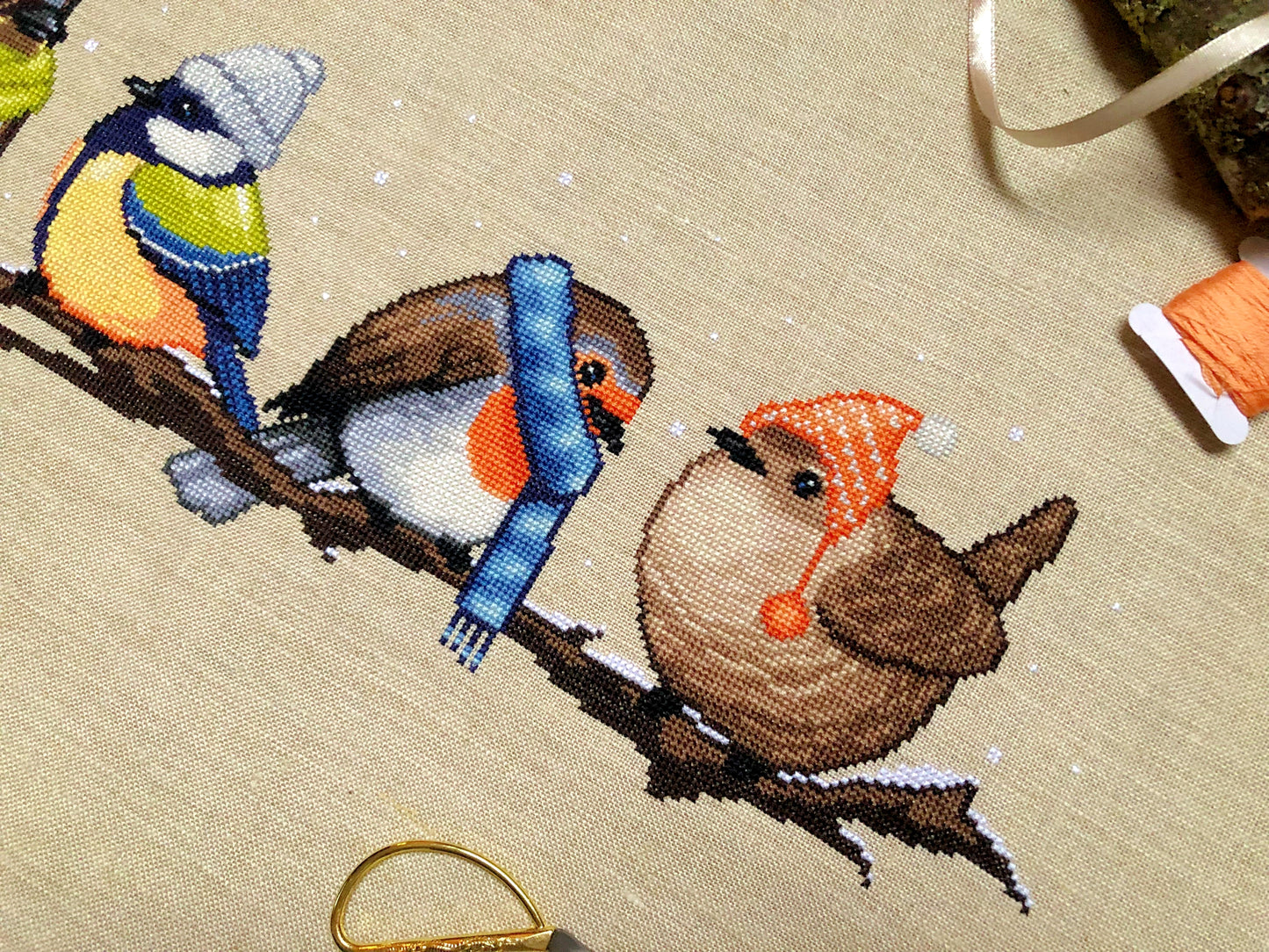 Closeup of Winter Birds cross stitch pattern. Two of the four birds are depicted, these are the robin and wren. Colors are yellow, brown, orange, blue, green and white. Robin is wearing a scarf and the wren is wearing a hat with dangly pompoms.