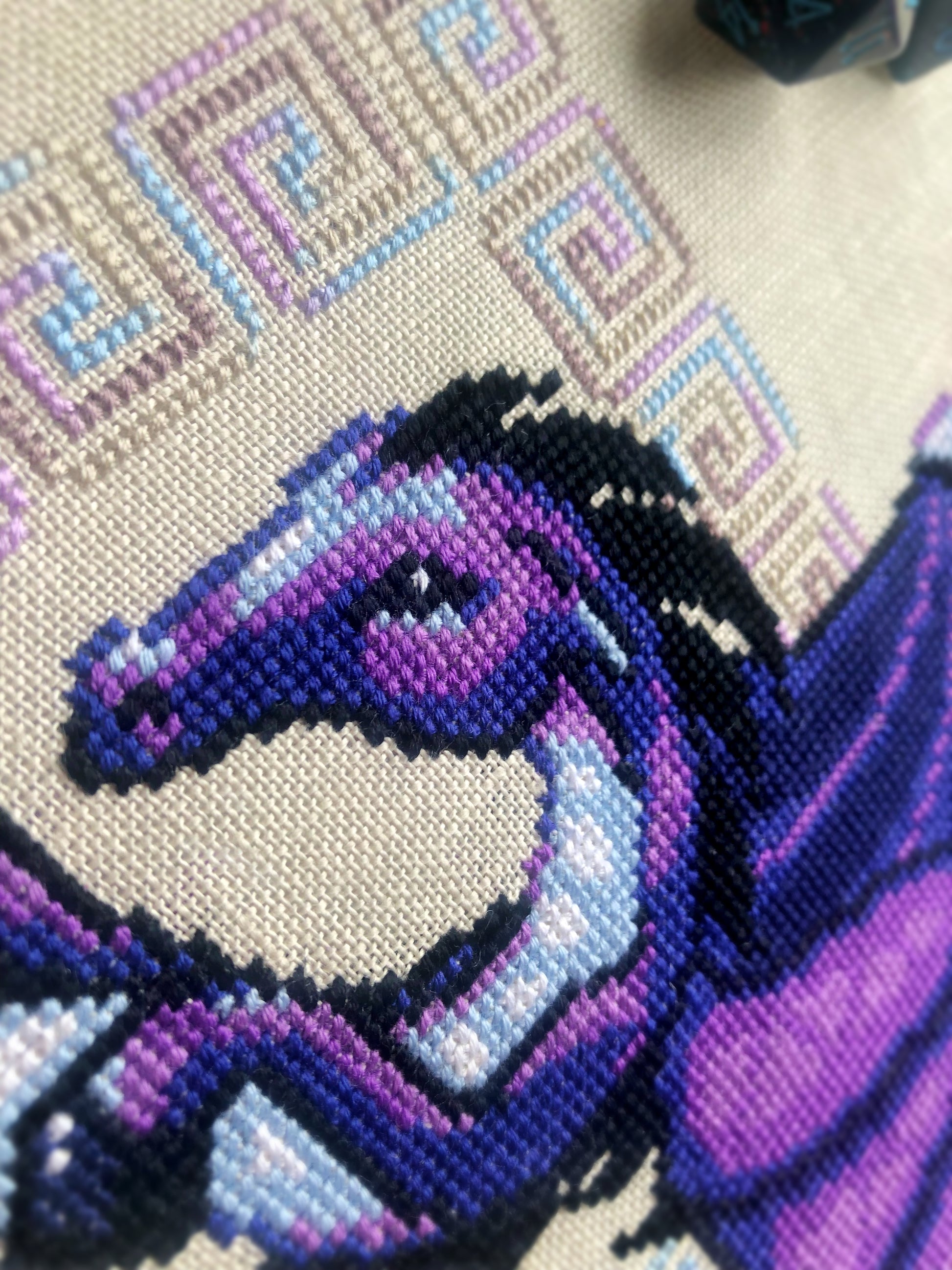 Closeup of Dragon cross stitch pattern. Stitched item, surrounded by decorations. Finished piece is of medium to large size. Colors are blue, purple, white, black and pink. Dragon is surrounded by variegated frame. Its head is like that of a horse.