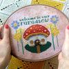Embroidery hoop held in hand, to show scale. Pattern is of medium size. Backstitches in the toadstool top are clearly visible here. Quote also contains backstitches and fractional stitches. Behind the scene is a crochet blanket in light colors.
