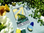 Flatlay of The Friend frog tarot cross stitch pattern. Stitched item, surrounded by decorations. Finished piece is of small to medium size. Colors are yellow, green, blue, grey and black. Frog is cute and small. The Friend is written underneath.