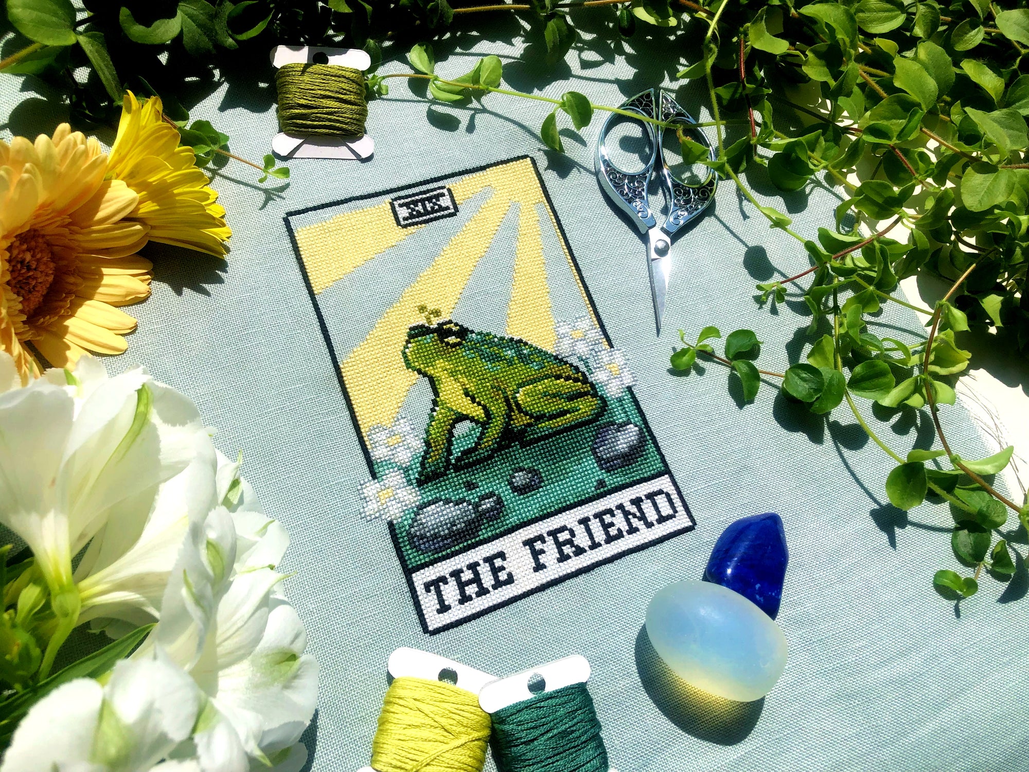 Flatlay of The Friend frog tarot cross stitch pattern. Stitched item, surrounded by decorations. Finished piece is of small to medium size. Colors are yellow, green, blue, grey and black. Frog is cute and small. The Friend is written underneath.