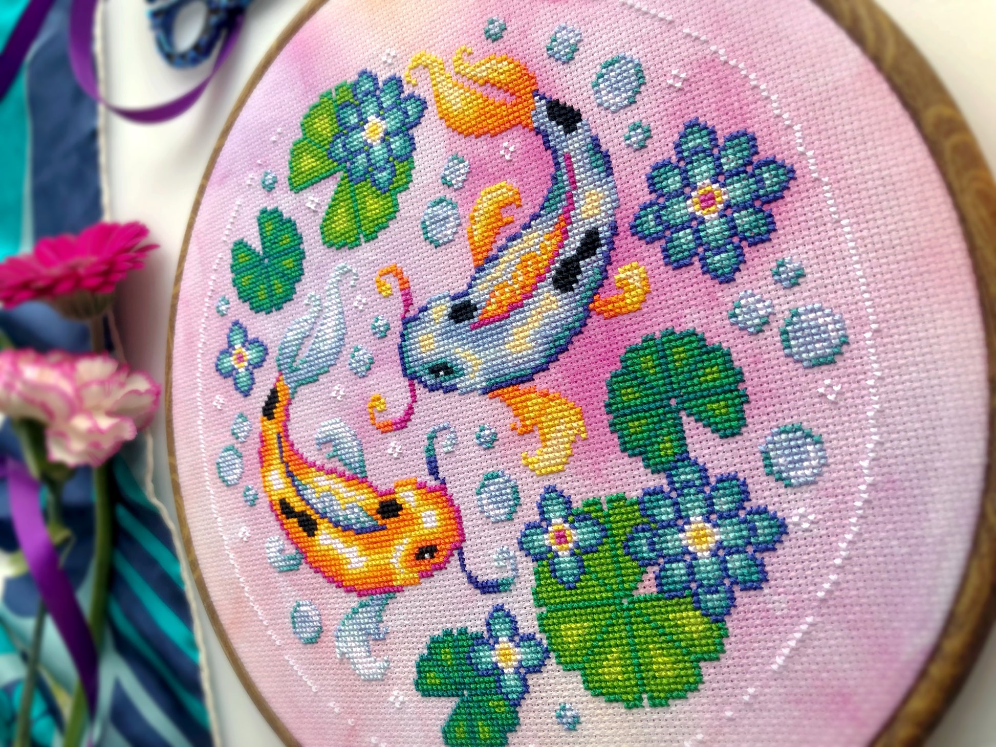 Closeup of Koi Fish cross stitch pattern. Right side is most visible. Finished piece is of medium size. Colors are very bright, and are green, blue, orange, pink and yellow. There are two fish, surrounded by lotus leaves and flowers.