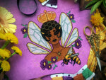 Front view of Queen Bee cross stitch pattern, slightly tilted. Colors are bright and vibrant. The piece is stitched on a warm purple 16ct Aida by EasyStitchForFun. Queen has a honeycomb pattern in her hair, and hair clips. The wings look delicate.