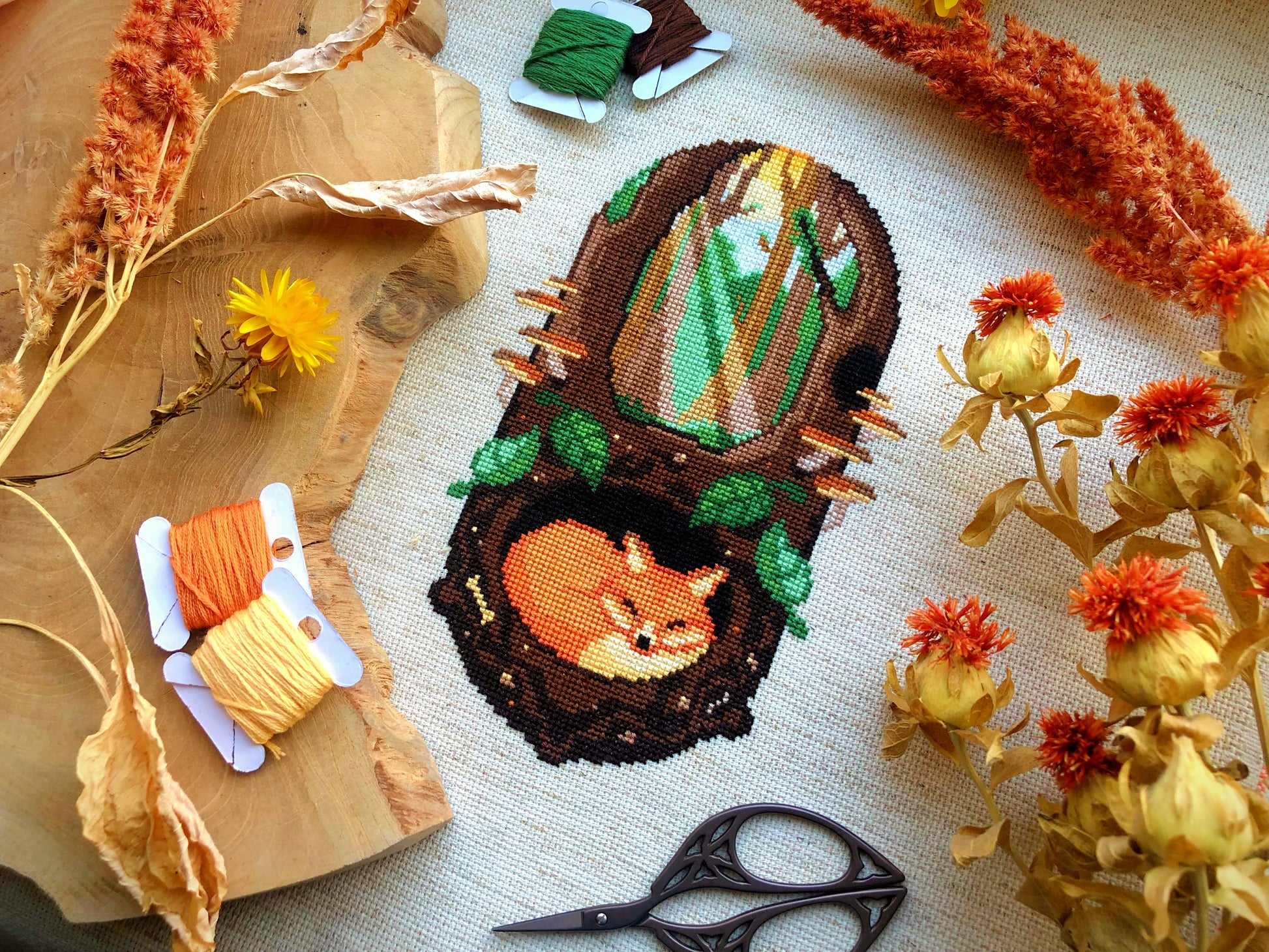 Flat-lay of Sleeping Fox cross stitch pattern. Stitched item, surrounded by decorations. Finished piece is of medium size. Colors are green, brown, orange, black, beige and white. Fox is sleeping underground. Trees and sun rays are above.