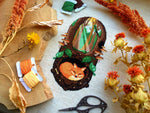 Flat-lay of Sleeping Fox cross stitch pattern. Stitched item, surrounded by decorations. Finished piece is of medium size. Colors are green, brown, orange, black, beige and white. Fox is sleeping underground. Trees and sun rays are above.