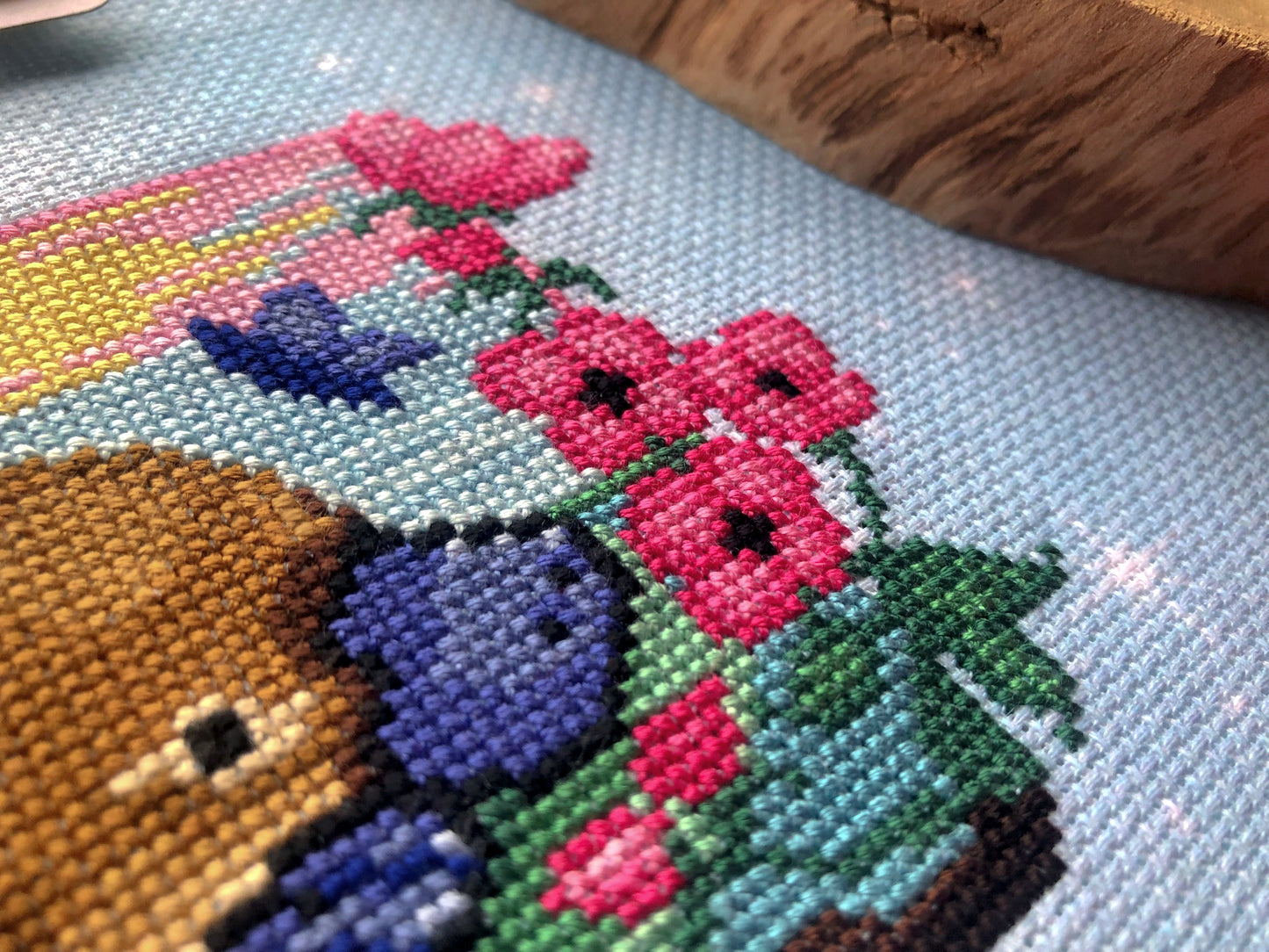 Extreme closeup of righthand side of platypus cross stitch pattern. Platypus is sniffing the pink flowers, which have black cores. Stitches are neat and tidy. Colors are vibrant. Platypus is adorable.