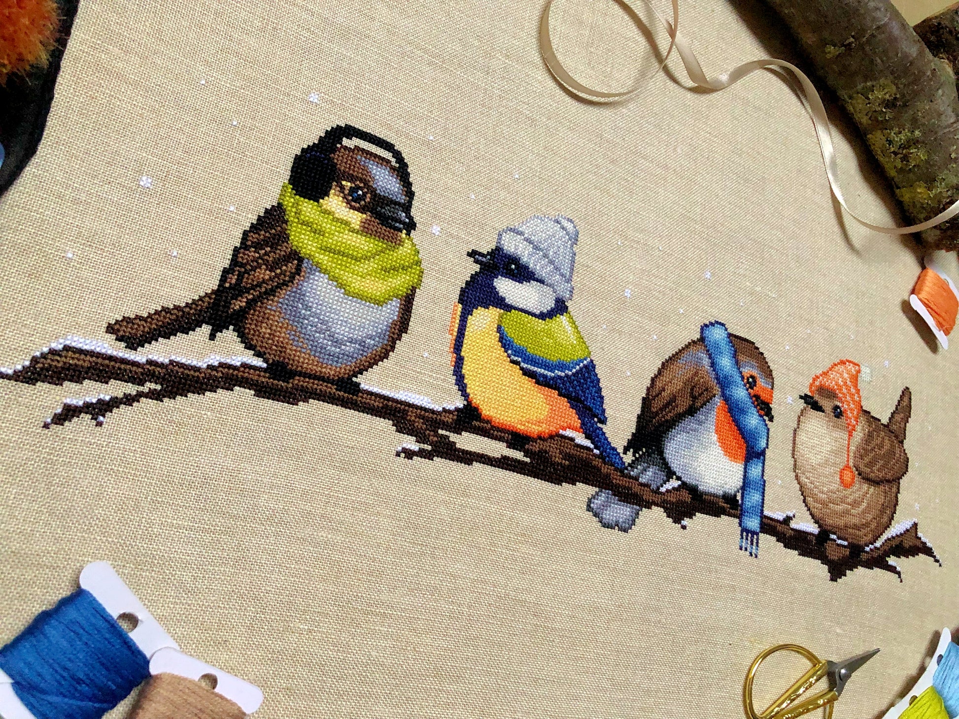 Flatlay of Winter Birds cross stitch pattern. Stitched item is surrounded by decorations. Finished piece is of medium to large size. Colors are yellow, brown, orange, blue, green, white. There are four birds sitting on a branch wearing winter gear.